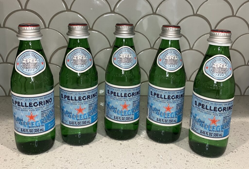 Five mini Pellegrino green glass water bottles aligned in front of a scalloped shell backsplash. Red stars are center on the bottles.